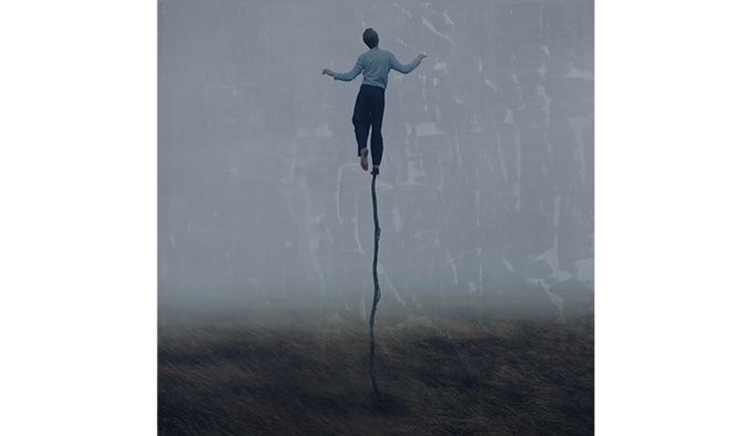 A  figure atop of a thin pole in the centre of a field. Their arms outstretched.  In the background the sky is grey.