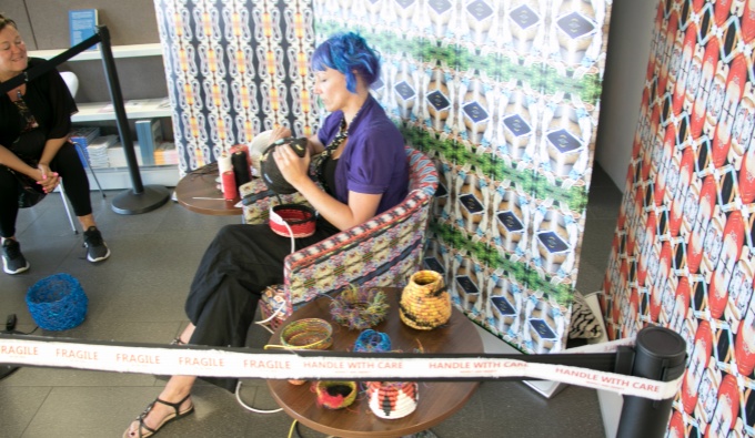 Kristina sits in a gallery space in a patterned tub chair with vibrant, patterned paneling behind her. She has blue hair, purple top and dark trousers. She weaves a basket behind a cordon that reads; fragile.