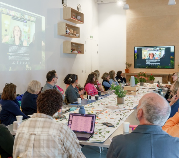 A cohort of over 18 arts professionals are seated around a long rectangular table in a brightly lit, white walled room with high ceilings. They are all looking towards a speaker attending remotely via zoom projected onto the wall.