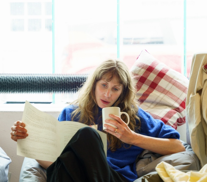 Nathalie is a white woman with long blonde hair and wears a bright blue jumper with rolled up sleeves, she is sat on a beanbag reading meeting information with cup in her hand.