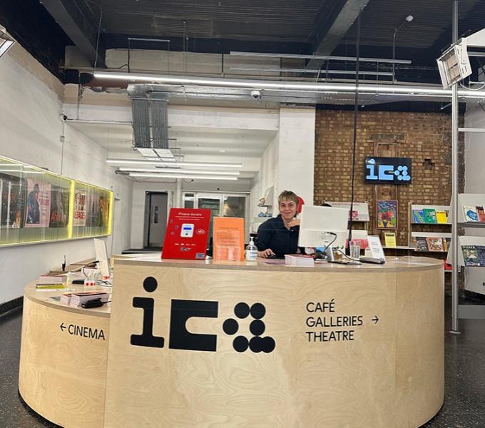 A large open space with a large curved wooden reception desk with a person seated behind it. There is a large screen on the desk as well as leaflets and notices. Across the wooden front are the organisation initials ICA. 