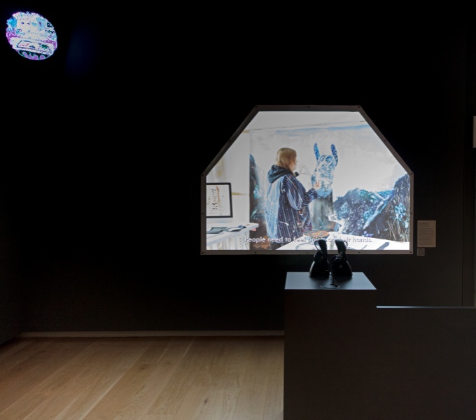 A dark gallery space with the film by artist Grace Currie projected in an arch shape onto the far gallery. There is a silouhette of black, Doc Martin style boots on a plinth in the foreground. 