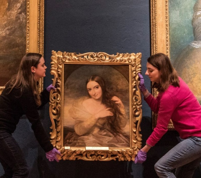 Two women hold a large gilt framed old oil painting. They are crouching as it to lift it up onto a navy blue gallery wall. Both wear white gloves. 