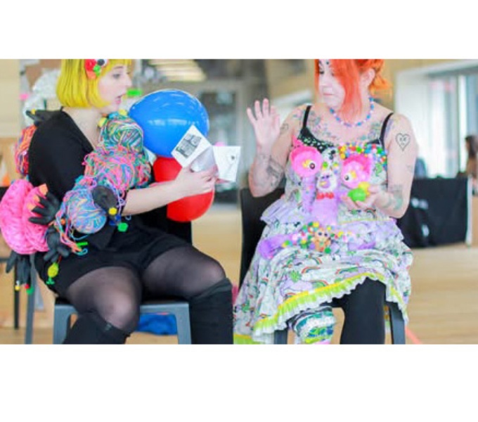 Image description: Two women seated in a large room. They both have vibrant coloured hair and wear brightly coloured clothes. The woman on the left holds out a folded paper fortune teller.