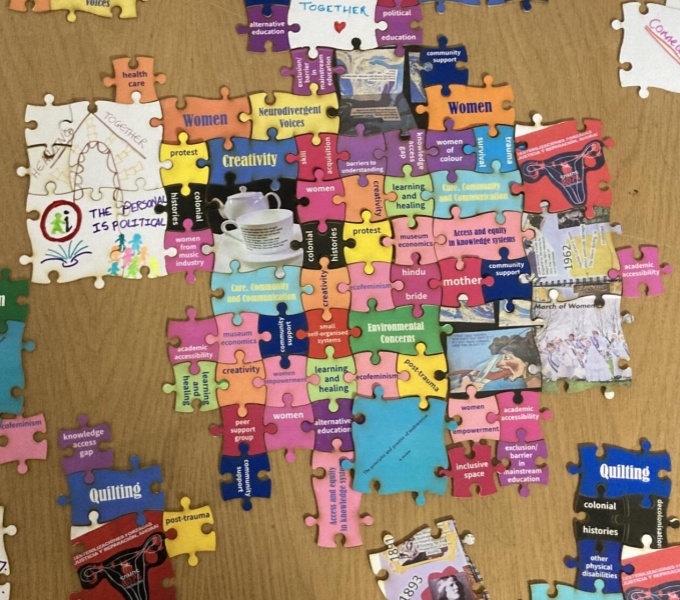 A view of a table top from above. Spread across the table are different coloured jigsaw pieces with short words or phrases on them. Including activism, quilting, learning and healing, creativity, colonial histories and academic accessibility.