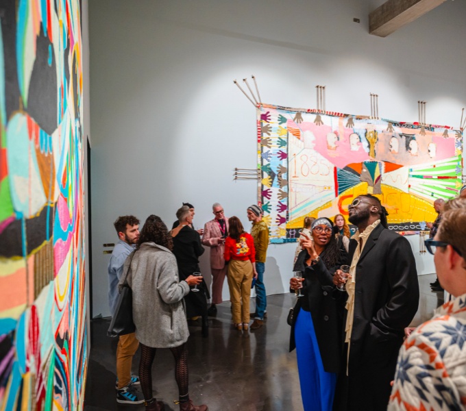 A crowded gallery space. To the left a large painting on the wall. Ahead and to the right people mingle and chat. To the centre tow people point and gaze at the artwork in discussion.