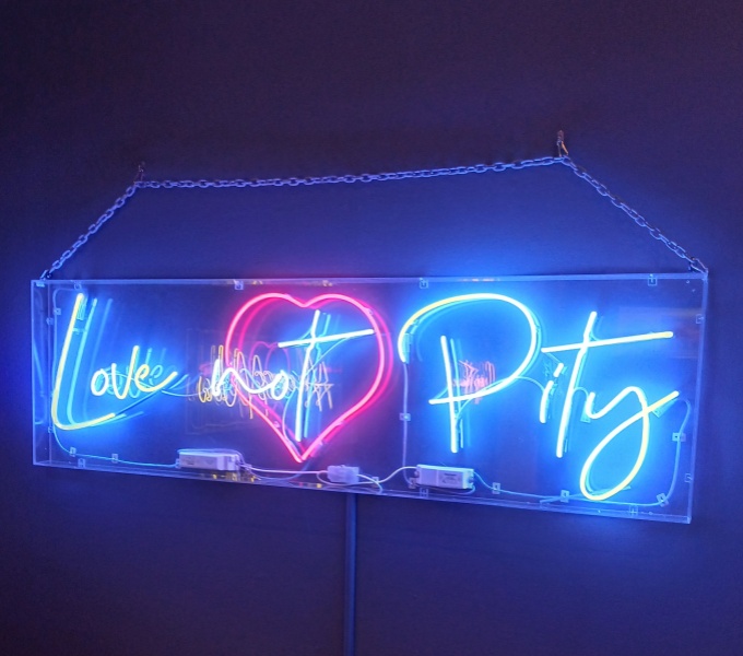 A neon sign of the words 'Love not Pity' in blue neon. Around the word 'not' is a neon red heart.