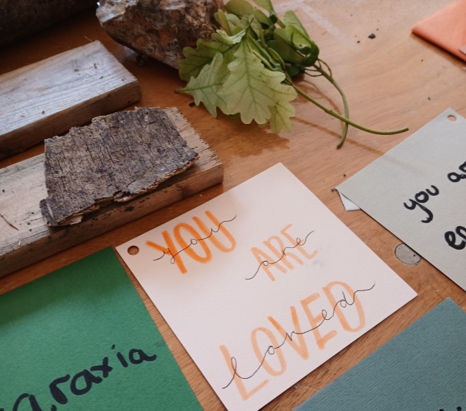 Am arrangement of young green oak leaves, small wooden logs and coloured papers on a table. In the centre is a pink square of card with the hand written message; You Are Loved.