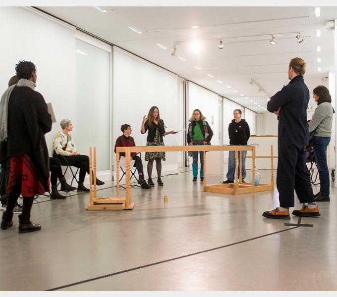 A crowd of people stand around a structure in the centre of a white gallery space. 