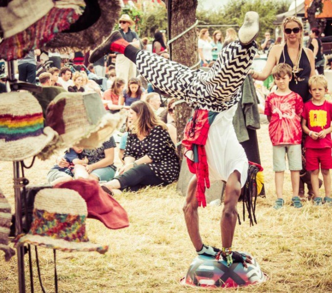 A crowd of festival goers, including young children gather around an acrobat doing a handstand. The acrobat wears black and white striped baggy trousers and a white tee-shirt. In the foreground to the left of the image is hat stand full of colourful hats.