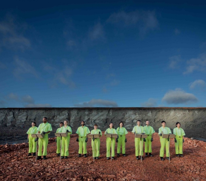 Twelve people wearing flourescent yellow trousers, shirts and gloves, are stood facing the viewer in an arid landscape. The sky above is blue.