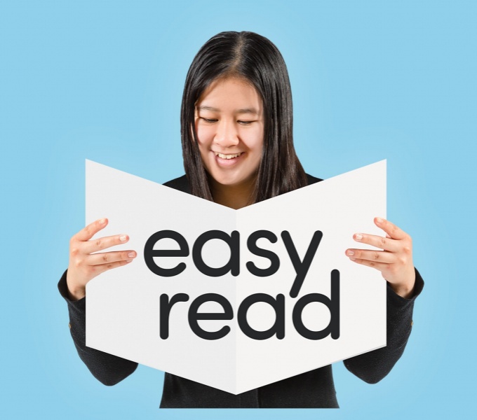 A n image of a young woman holding a booklet with the words easy read across it, open in front of her. She wears a black top and has long dark hair and is stood against a pale blue background.