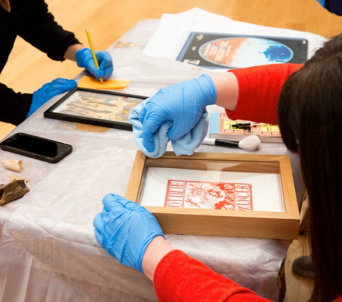 A table with assorted framed artworks across it. Either side are two people wearing gloves working. One is cleaning a framed work the other writes notes on a yellow piece of paper.