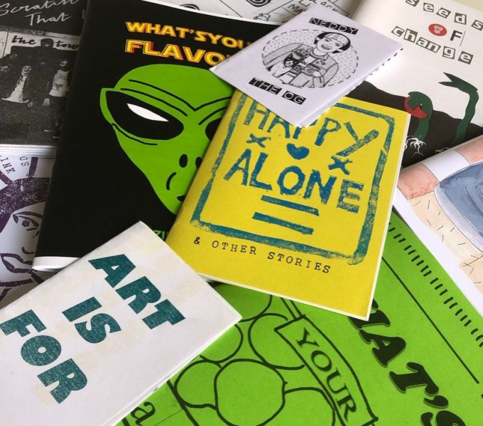 A selection of Zines spread out and overlapping. To the bottom left is a zine cover in white with the words Art is For, printed in green ink. Other titles include, Happy Alone, Seeds of Change and What's Your Flavour? 