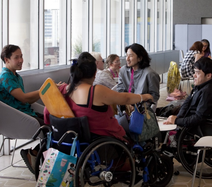 A group of young adults sit talking around a low table in a well lit glazed room. The person in the foreground with their back to the camera is sitting in a wheelchair.