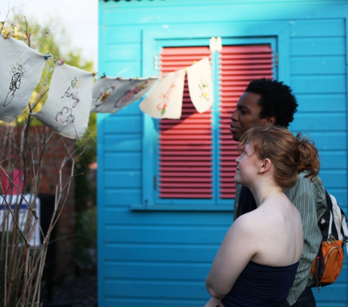 A young black man and young white woman stand in front of a bright blue wooden building with red shutters. Facing to the left they look at art works, hung on string, blowing in the breeze. It's a beautiful day, the sky is blue and the sun is shining.
