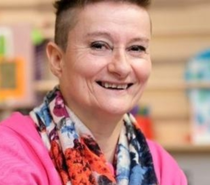 Artistic Director Tanya wears a pink cardigan and patterned shirt. She has short cropped hair and smiles as she looks at the camera. 