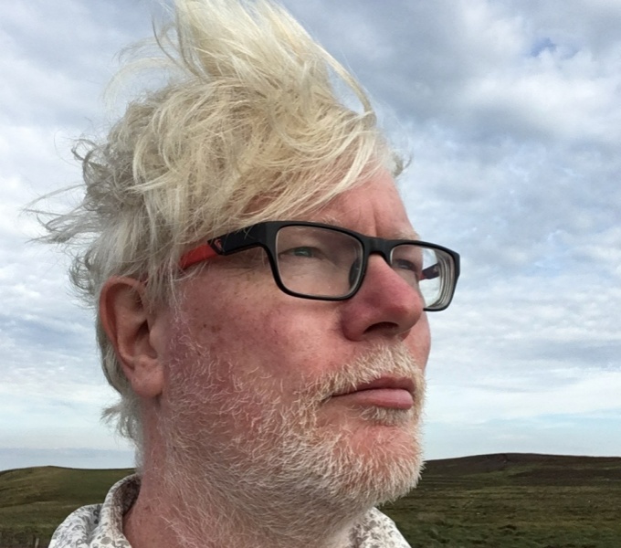 A head shot photo of Aidan Moesby. He stands against a blue, cloudy and stormy looking sky, his white blonde hair blowing backwards in the wind. He wears dark rimmed glasses and a pensive expression on his face.