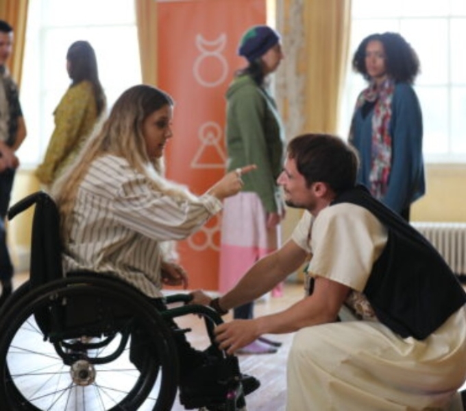 A woman seated in a wheelchair points towards a man kneeling in front of her. He looks at her directly as she speaks. In the background are several onlookers.