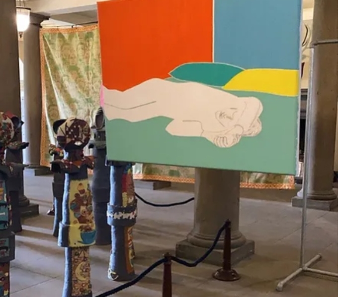 Image shows a brightly coloure canvas and floor standing totem pole style sculptures on display in a room with pillars and high ornate ceilings.