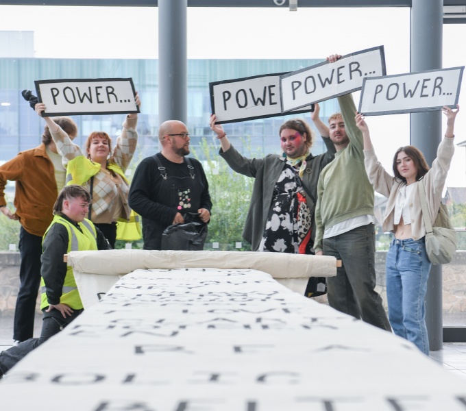 Seven people stand at the foot of a long narrow table. A large scroll in un-raveled upon it. Two of the people wear hi-vis tabards and four are holding placards that say, POWER, above their heads. They stand in front of a large glass window.