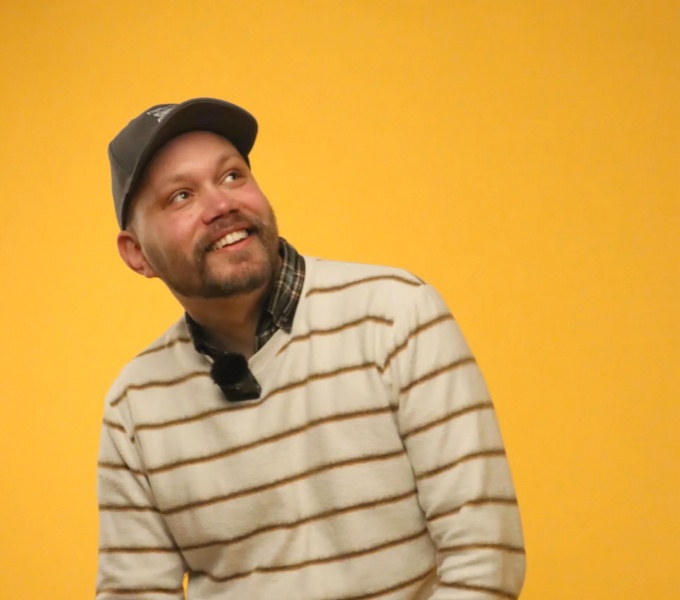 A man in a cream and brown striped jumper and flat cap is seated against a deep yellow background. He has facial hair and gazes smiling towards the right hand side of the image.