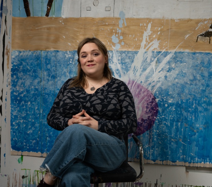 A portrait photograph showing a white woman with long brown hair sitting in a low swivel chair in front of a painting entitled Grace and John’s Biggest Splash! 