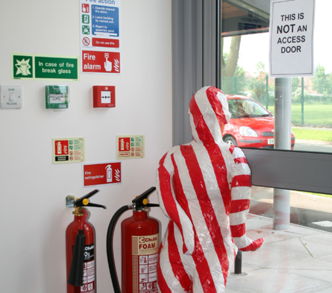 A figure like form wrapped in red and white tape stands in front of a glass door with a sign in capital letters saying this is not an access door