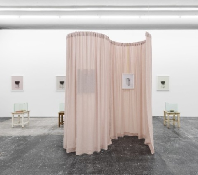 A white gallery space. In the centre hangs a floor to ceiling cream curtain, that hangs in an S shape. On the far wall are framed artworks.