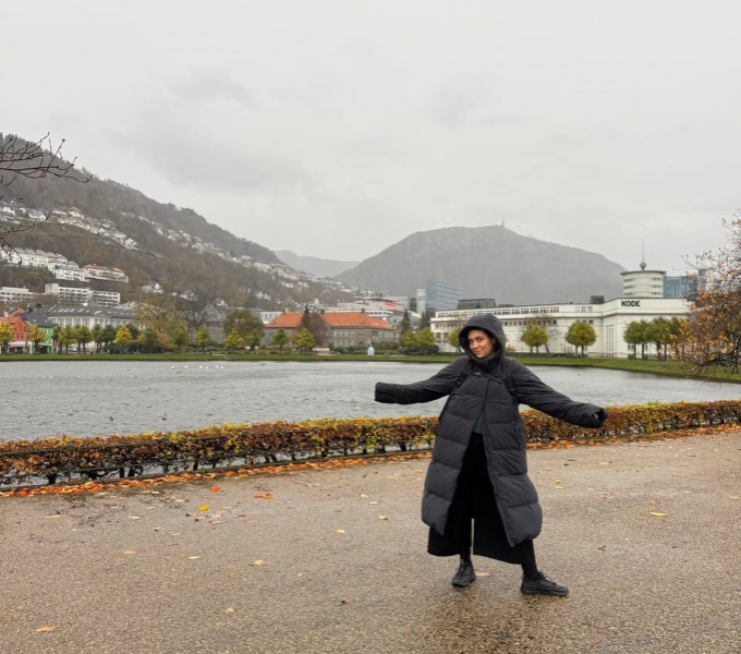 A woman in a long black padded coat is stood in front of a body of water. Mountains and buildings are in the distance set against a dark grey sky.