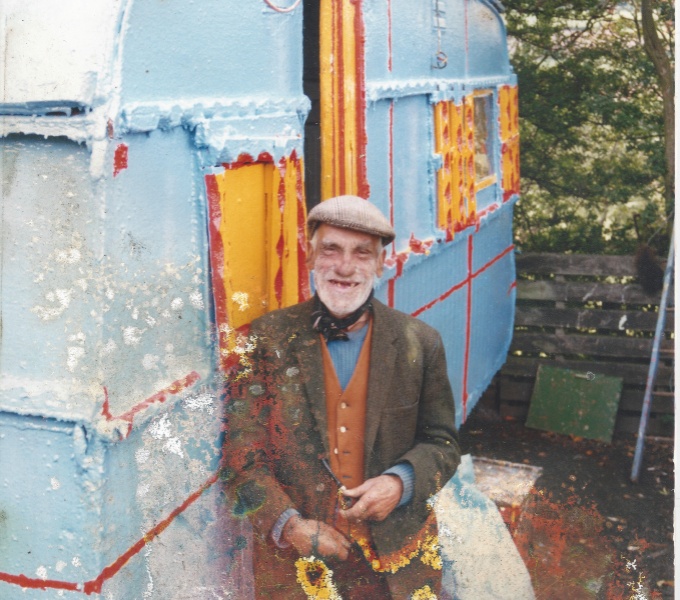 Bill locke stands in front of a blue tourer caravan with yellow and red shutters. He wears a flat cap, a scarf tied around his neck, with a blue jumper, orange waistcoat and brown jacket. He grins broadly at the camera. 