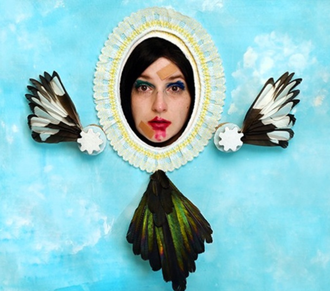 A face with smeared make up protrudes through a hole in a painted blue backdrop decorated with feathers