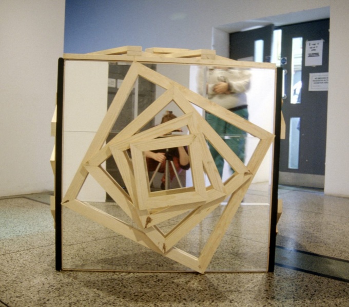 A mirrored square sculpture in a large room. Attached the face of the mirrored square facing the camera are several wooden square frames attached, every decreasing in size and leaving a small square at the centre of the mirror exposed.