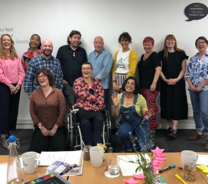 DASH staff and trustees lined up against a white wall in two rows, smiling and pulling silly faces at the camera. in the foreground is a table littered with papers, pens and forget me nots.