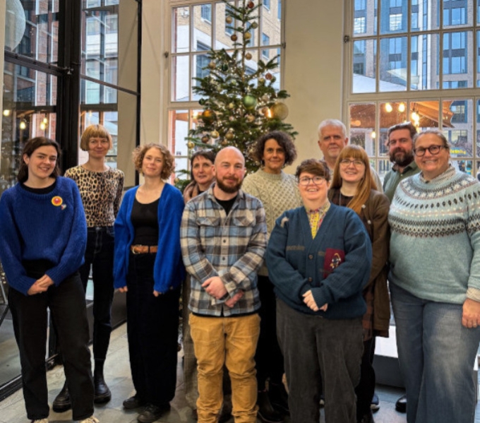 Members of the DASh team and board stand in front of a Christmas tree in the glazed foyer of IKON gallery