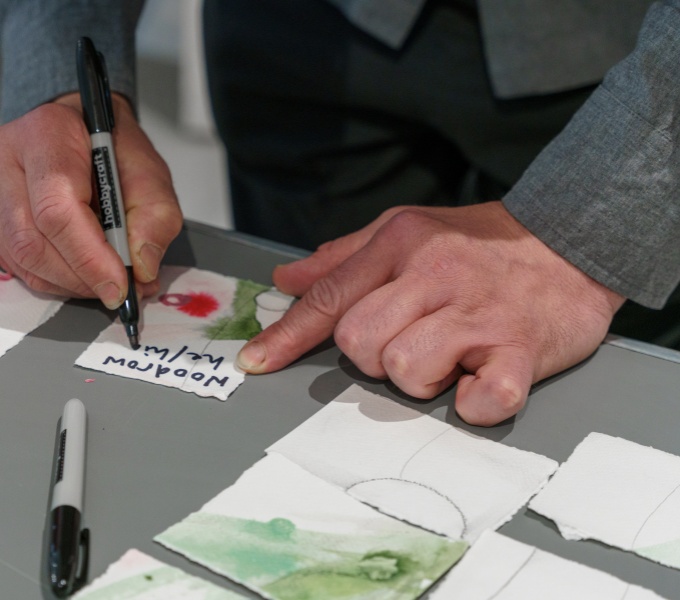 Hands rest on a table. The right hand writes a name badge in black marker on water colour paper decorated in similar design to the table centre piece.