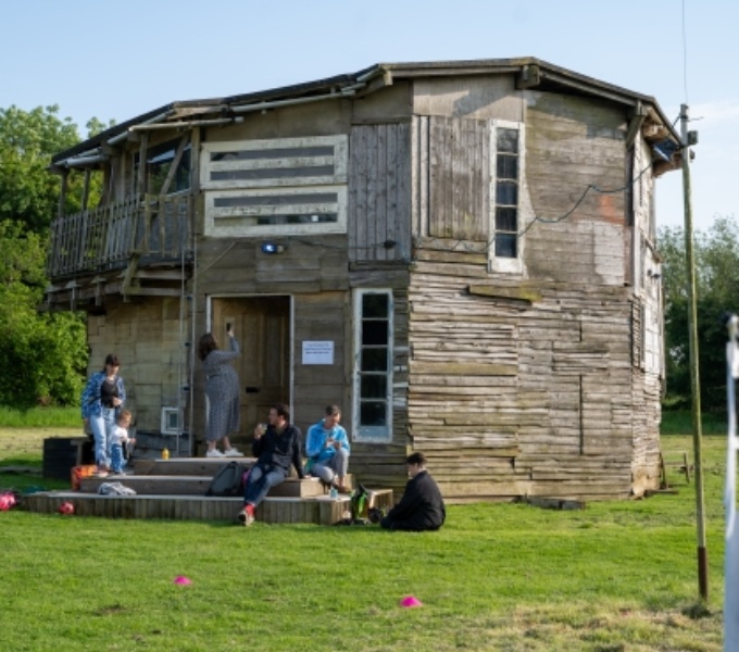 People are congregated outside of a large handmade building made of recycled wood, windows and doors. It is situated in a green field with trees in the distance. Its a pleasant day with blue but slightly cloudy skies.