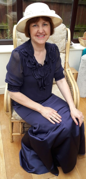 Image shows Processions participant Edel Slawson sitting in a chair, facing the camera and smiling. She is wearing a full length purple satin dress in an early 20th century style, with a cream hat with flowers.