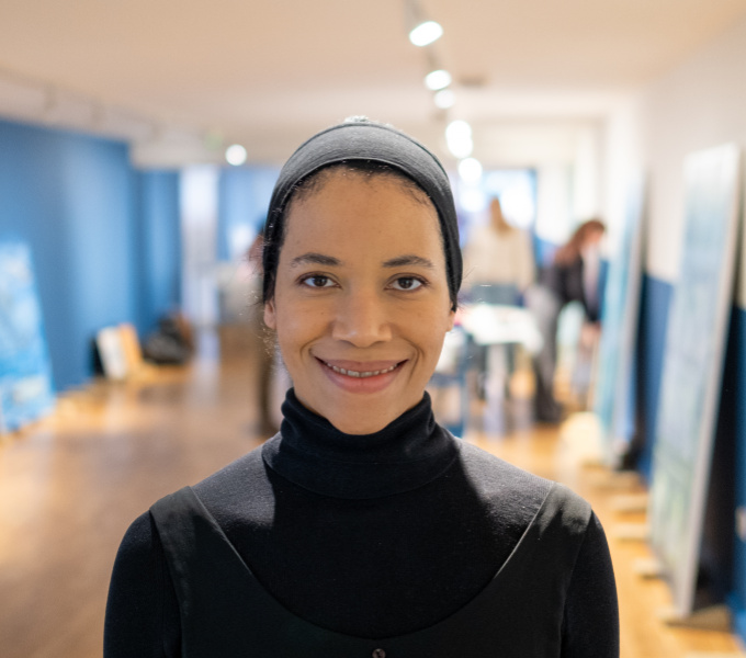 Eve stands in a gallary space with blue walls and light brown floor. Paintings lean up against the walls behind her. She faces the camera, wearing her hair pushed back in a wide headband, and a black, roll-necked top.