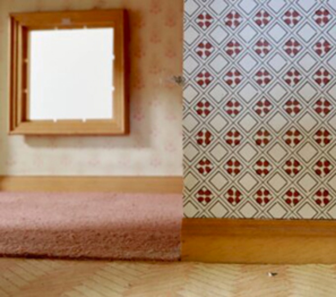 A room of two halves. To the right s a wall covered in bold retro print wallpaper in red and black. To the left is a large square mirror mounted on the wall. Below is a raised floor, covered in a thick piled pink carpet. 