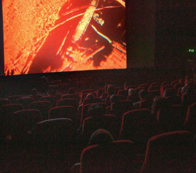 A dark cinema room with tiered seating. On the large screen is an un-identifiable image in orange and black.