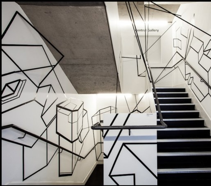 Image description: A stairwell with black steps and handrail. The walls alongside are white with angular shapes marked out in thick black lines.