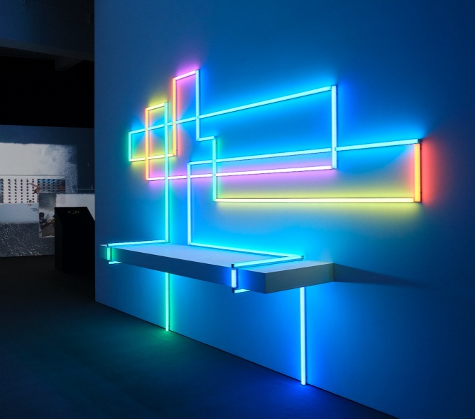 A dark gallery space. On the wall immediately ahead is a large artwork made of florescent tube lighting. A series of horizontal tubes are lit up in electric blue and bright yellow, whilst the adjoining vertical tubes to the sides glow pink and red.