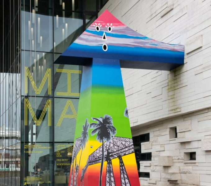 A large sculpture stands outside the large glass windows of MIMA. It is almost mushroom in shape. It is painted in rainbow colours with a face painted on the triangle at the top. 