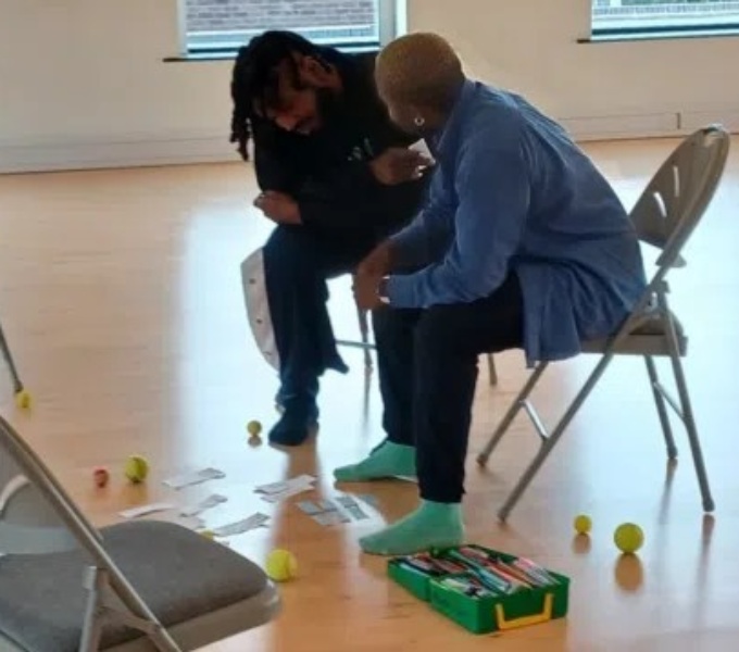 Two people sat on chairs talk to each other as they look at paper and balls scattered on the floor. 