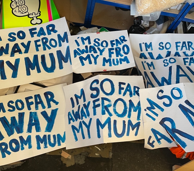 Image shows six hand painted banners with blue capital letters on white paper, which read 'I'm so far away from my mum'.
