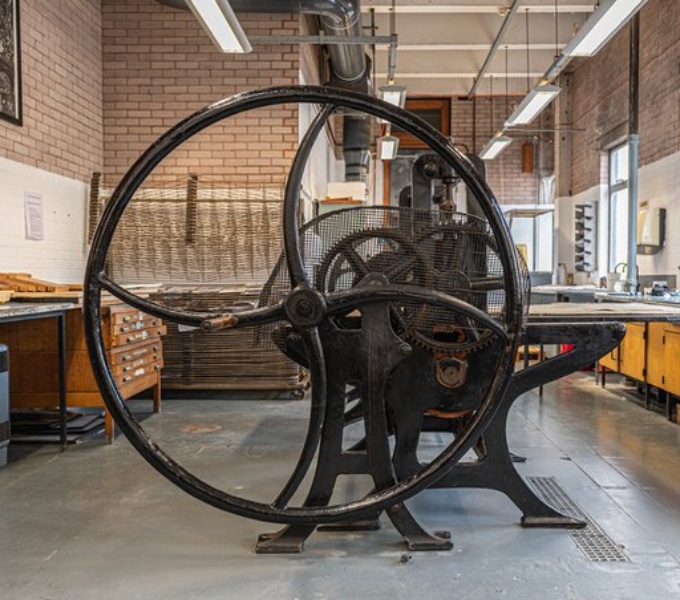A large workspace with a traditional printing press with large fly wheel at the centre. 