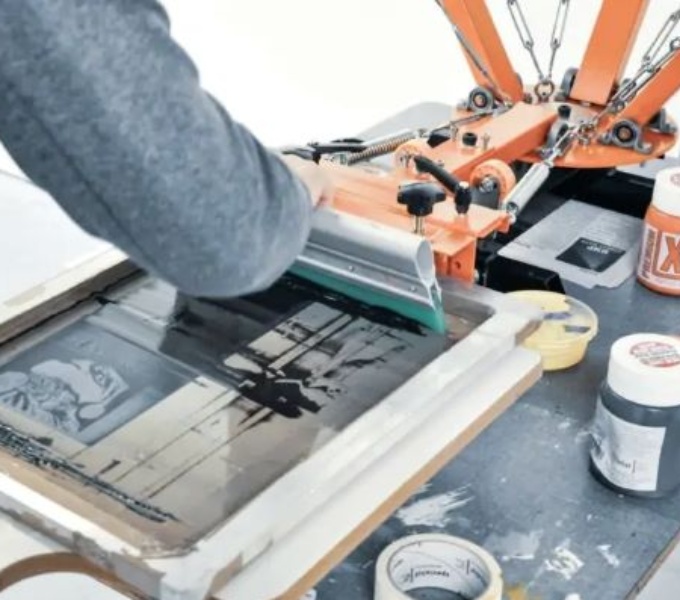 Printing equipment and a screen on a table in a white room. An arm is in view, about to pull the ink over a screen with a squeegee.