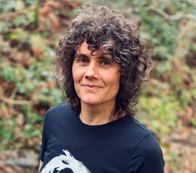 Heather has brown shoulder length curly hair. She faces the camera and wears a navy blue t-shirt with  a circular printed design in white. She stands in her own woodland, part of the Celtic Rainforest Wales rewilding program. 