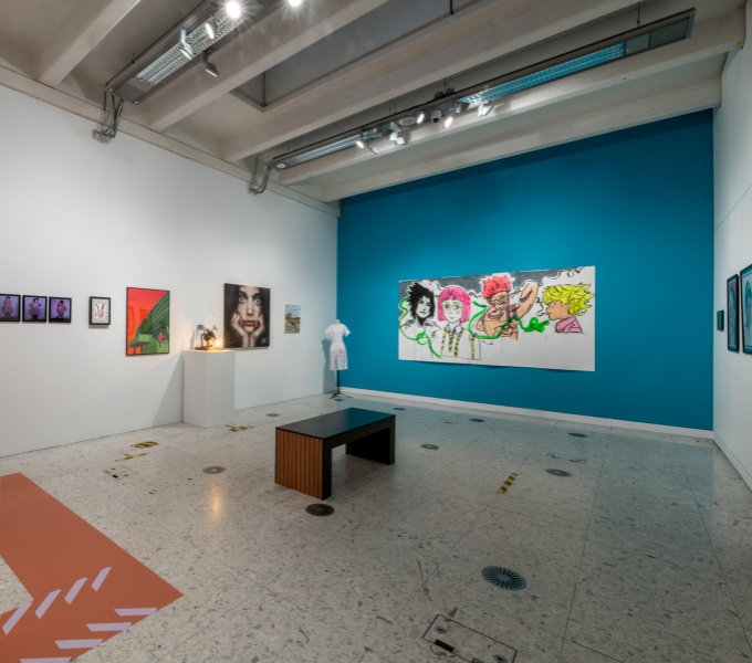 A large gallery space with two white walls to the sides, with far wall painted a turquoise blue. Assorted artworks are hung  on the walls. In the centre of the space is a simple wooden bench, as well as a white table and chairs.